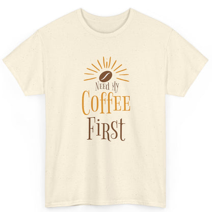Beige t-shirt with 'Need My Coffee First' text and coffee bean graphic on a white background