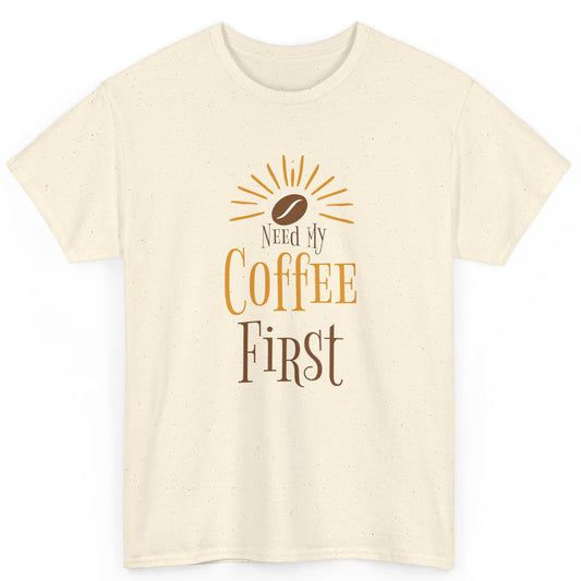 Beige t-shirt with 'Need My Coffee First' text and coffee bean graphic on a white background